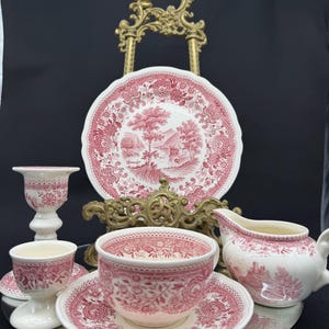 Può includere: Una collezione di stoviglie vintage in ceramica rosa e bianca, tra cui un piatto, una tazza e un piattino, una crema, un portauovo e un candelabro. I pezzi presentano un design floreale e paesaggistico dettagliato. Gli articoli sono disposti su una superficie a specchio.