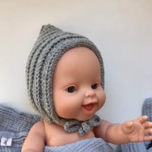 Könnte beinhalten: Eine graue gestrickte Babyhaube mit einer spitzen Spitze und einer Schleife am unteren Ende. Die Haube befindet sich auf einer Baby Puppe mit einer hellblauen Decke.