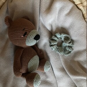 May include: A brown crocheted teddy bear with light green accents sits on a light gray knitted blanket. A light green crocheted flower is also on the blanket.