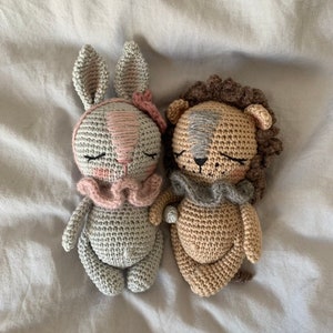 May include: A gray crocheted bunny and a brown crocheted lion laying on a white bedspread. Both stuffed animals have closed eyes and are wearing pink and gray crocheted collars.