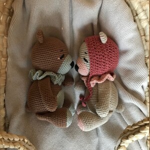 May include: Two crocheted teddy bears, one brown with a green scarf and one beige with a pink hat, are lying on a light gray knitted blanket inside a woven basket.