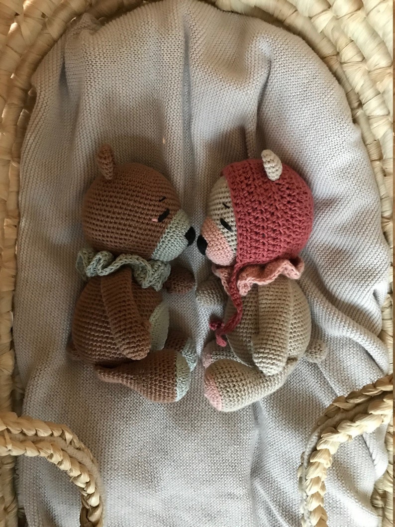 May include: Two crocheted teddy bears, one brown with a green scarf and one beige with a pink hat, are lying on a light gray knitted blanket inside a woven basket.
