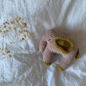 May include: A crocheted elephant toy with a pink body and yellow ears. The toy is lying on a white bedspread.