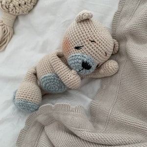 May include: A crocheted stuffed animal bear in beige and blue colors, lying on a beige knitted blanket with ruffled edges.