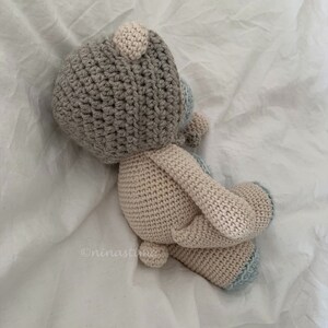 May include: A crocheted stuffed animal in the shape of a bear. The bear is light brown with a gray head and blue accents. The bear is lying on a white surface.