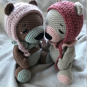 May include: Two crocheted teddy bears wearing pink and red hats. The bears are sitting on a white blanket and are facing each other.