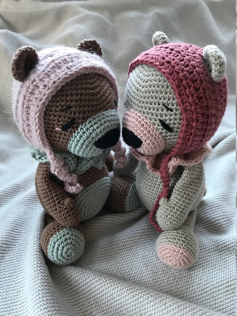 May include: Two crocheted teddy bears wearing pink and red hats. The bears are sitting on a white blanket and are facing each other.