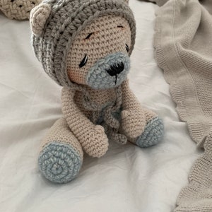 May include: A crocheted teddy bear wearing a crocheted hat with blue accents. The bear is sitting on a white surface.