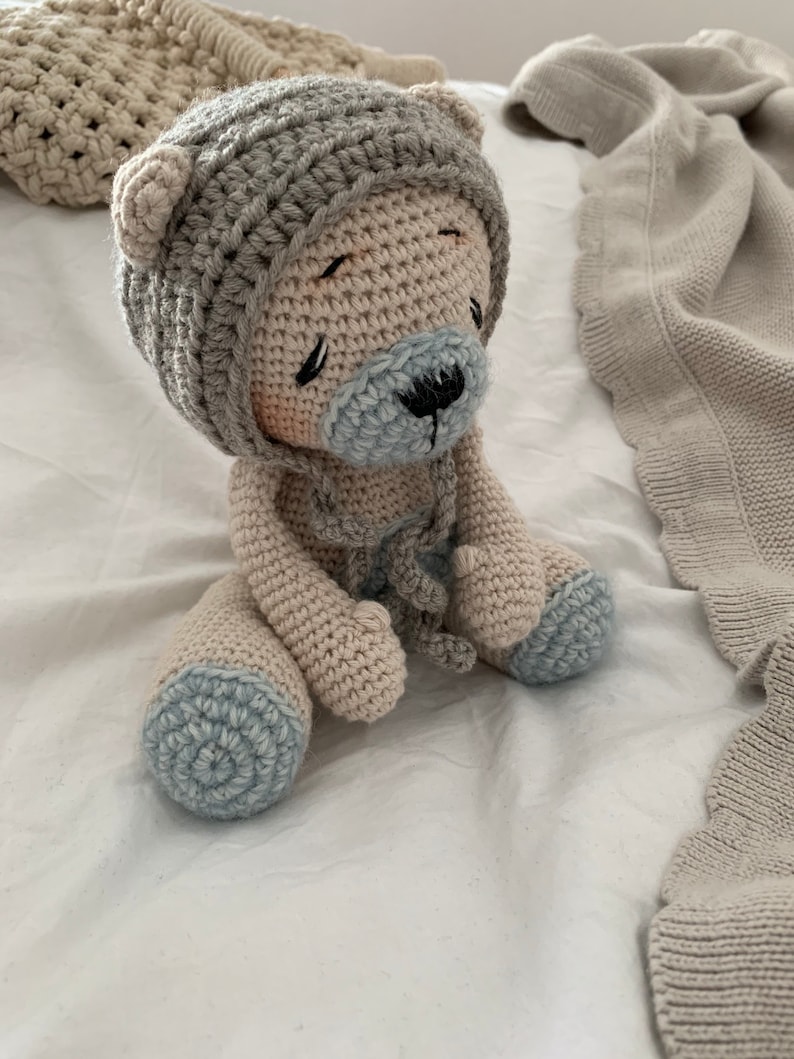 May include: A crocheted teddy bear wearing a crocheted hat with blue accents. The bear is sitting on a white surface.