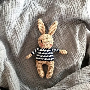 May include: A crocheted bunny toy with a striped shirt, lying on a gray textured blanket.