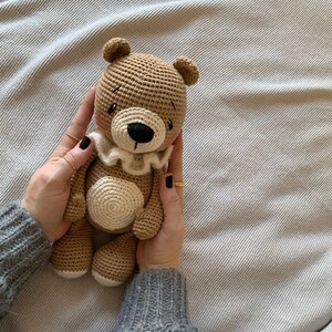 May include: A crocheted brown teddy bear with white accents, wearing a white ruffle collar. The bear is being held by hands with black nail polish.