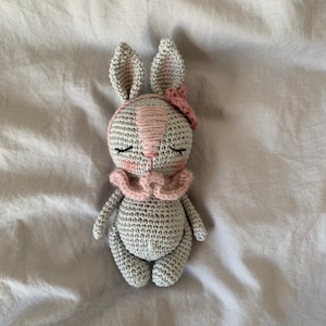 May include: A crocheted gray bunny with pink ears and a pink flower on its head. The bunny has closed eyes and a pink bow around its neck.