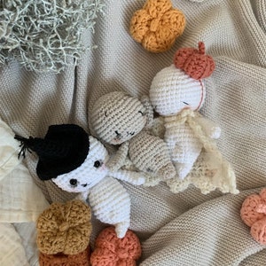 May include: A collection of crocheted amigurumi toys, including a ghost wearing a black witch hat, a small white ghost, and two beige ghosts. The toys are arranged on a beige knitted blanket with several crocheted orange and peach pumpkins.