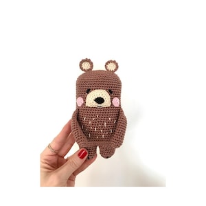May include: A brown crocheted bear with pink cheeks and a black nose. The bear has small, beige ears and is standing on a white background.
