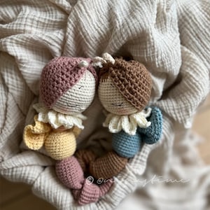 Może przedstawiać: Dwie połączone sznurkiem, szydełkowe lalki. Lalki mają twarze z brązowej i różowej przędzy z białymi akcentami z przędzy. Lalki noszą szydełkowe stroje w kolorze żółtym i niebieskim.