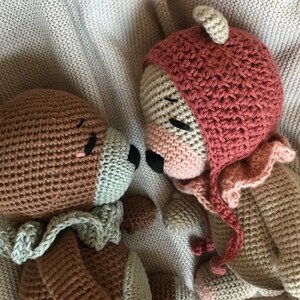 May include: Two crocheted stuffed animals, a brown bear with a green scarf and a tan bear wearing a pink hat, are lying next to each other on a beige blanket.