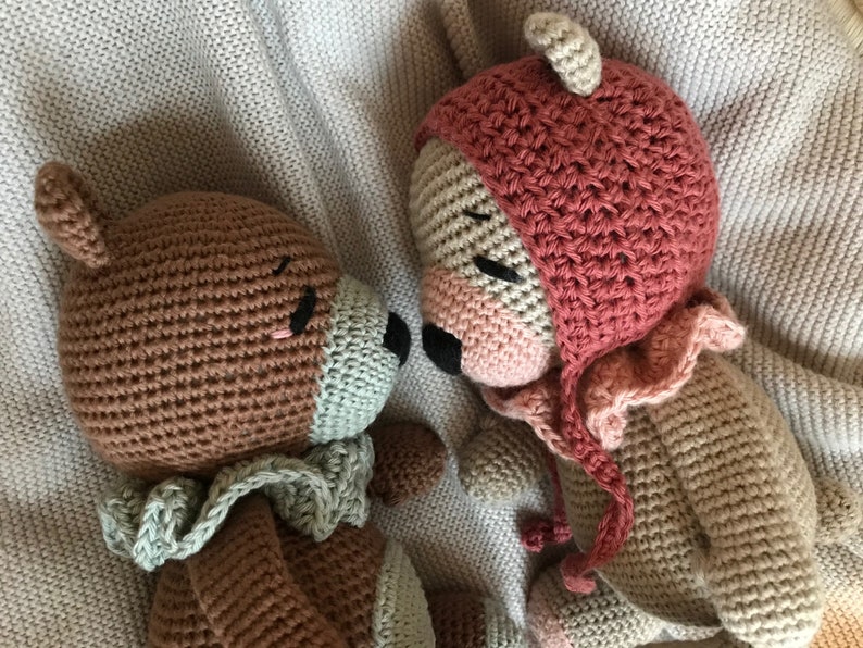 May include: Two crocheted stuffed animals, a brown bear with a green scarf and a tan bear wearing a pink hat, are lying next to each other on a beige blanket.