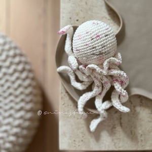 May include: A white crocheted octopus with pink accents. The octopus has a round head with embroidered eyes and a ruffled collar. The tentacles are long and curled.
