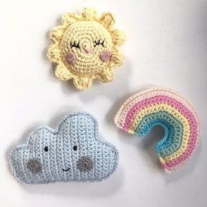 Könnte beinhalten: Drei gehäkelte Artikel: eine lächelnde Sonne, eine hellblaue Wolke und ein Regenbogen in Pastellfarben. Die Sonne ist gelb mit rosa Wangen, die Wolke hat graue Akzente und der Regenbogen hat rosa, gelbe und blaue Streifen.