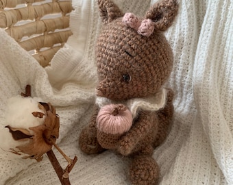 Könnte beinhalten: Ein braunes Häkel-Eichhörnchen-Spielzeug, das einen rosa gehäkelten Kürbis hält. Das Eichhörnchen trägt eine rosa Schleife und hat schwarze Augen. Das Spielzeug sitzt auf einer weißen Strickdecke.