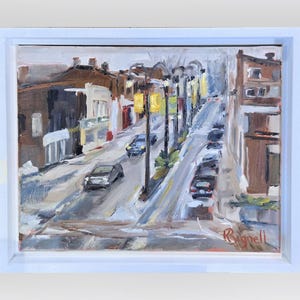 May include: Oil painting of a city street scene with buildings, cars, and streetlights. The artwork is framed in a white border. The painting uses a palette of browns, yellows, and grays to depict the urban landscape.