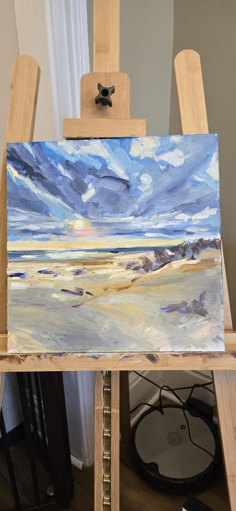 May include: An oil painting on a square canvas depicting a coastal landscape with a sandy beach in the foreground, a dark blue ocean horizon, and a dramatic sky filled with thick, textured brushstrokes of blue, white, and yellow clouds.