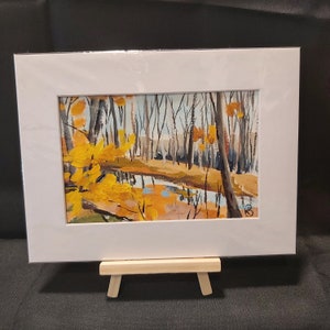 May include: A framed painting of a forest scene with a small stream in the foreground. The trees are bare and the leaves are a vibrant yellow. The painting is on a small easel.