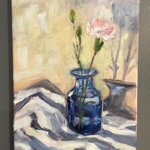 May include: An oil painting depicts a still life with a blue vase holding a pink carnation and green stems. The vase sits on a white and gray patterned cloth. A second blue vase is in the background. The background is a light yellow.