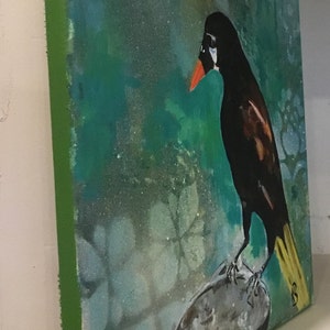 Op de afbeelding: Een schilderij van een zwarte vogel met een gele staart die op een grijze rots zit. De achtergrond is turquoise groen met een wit bloemenpatroon.