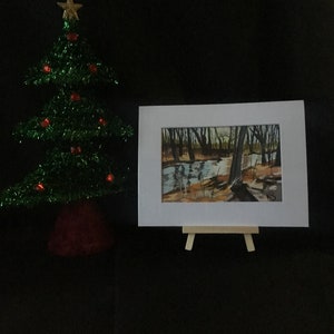 May include: A framed watercolor painting of a forest scene with a stream running through it. The painting is on a small easel and is in front of a green Christmas tree.