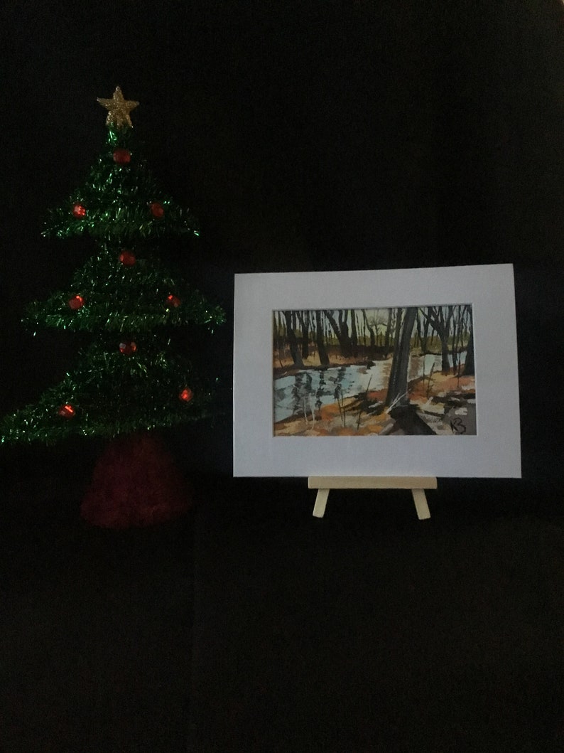 May include: A framed watercolor painting of a forest scene with a stream running through it. The painting is on a small easel and is in front of a green Christmas tree.