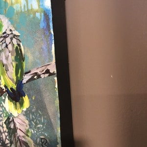 Puede incluir: Una pintura de un loro verde y blanco posado en una rama. El fondo es un dise&ntilde;o abstracto azul y verde con acentos blancos. La pintura est&aacute; enmarcada con un marco negro.