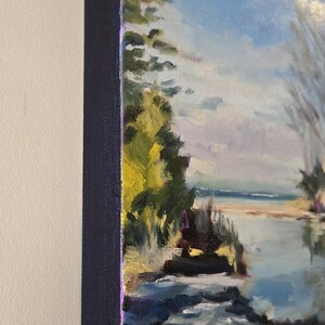May include: A painting of a landscape with a body of water in the foreground. The water is a light blue color and there are rocks and green grass in the foreground. The background is a light blue sky with white clouds. There are trees on the left side of the painting.