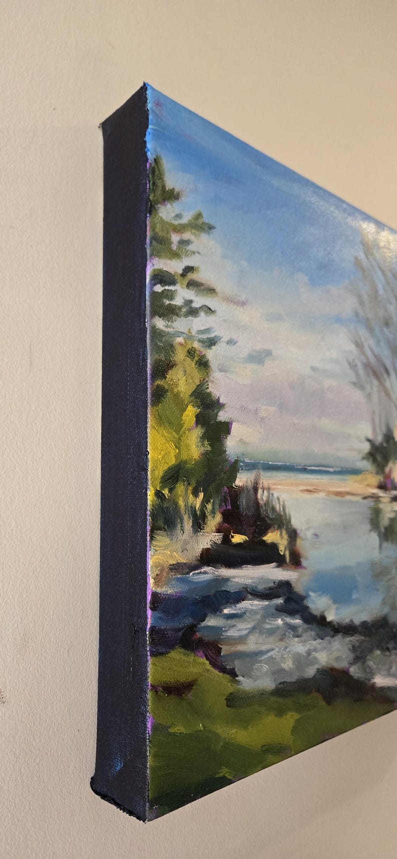 May include: A painting of a landscape with a body of water in the foreground. The water is a light blue color and there are rocks and green grass in the foreground. The background is a light blue sky with white clouds. There are trees on the left side of the painting.