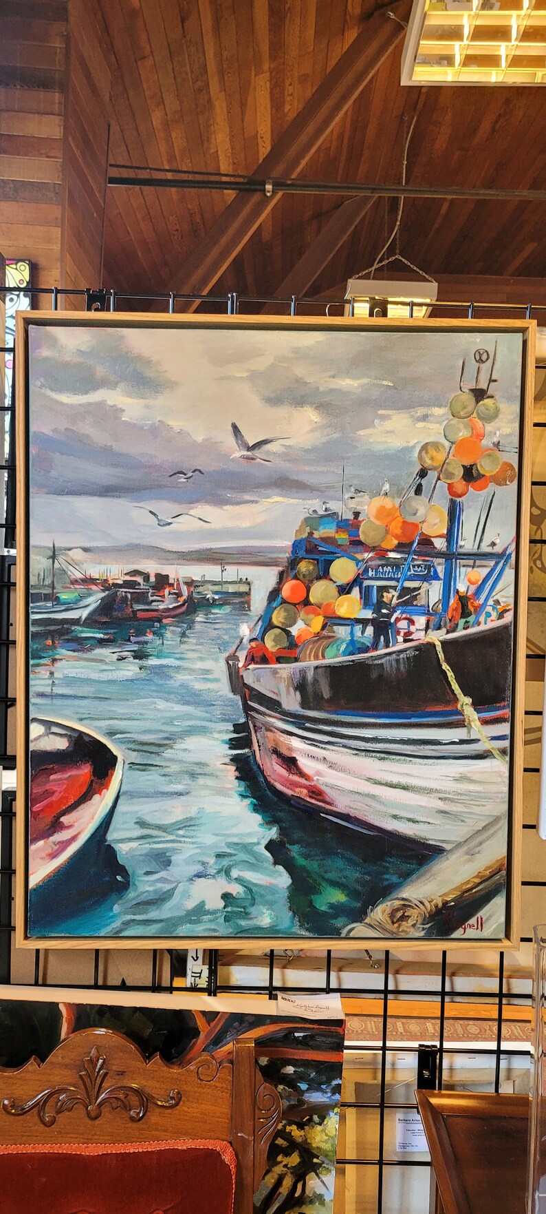 Op de afbeelding: Olieverfschilderij van een haven met vissersboten. Het kunstwerk toont een donkere boot met oranje en gele boeien, zeemeeuwen en een bewolkte lucht. Het schilderij is ingelijst in een licht houten frame.