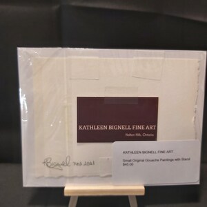 May include: A white card with a brown business card that reads "KATHLEEN BIGNELL FINE ART" and "Halton Hills, Ontario".  The card is signed "Bignell nov 2021".  A second white card reads "KATHLEEN BIGNELL FINE ART" and "Small Original Gouache Paintings with Stand $45.00".