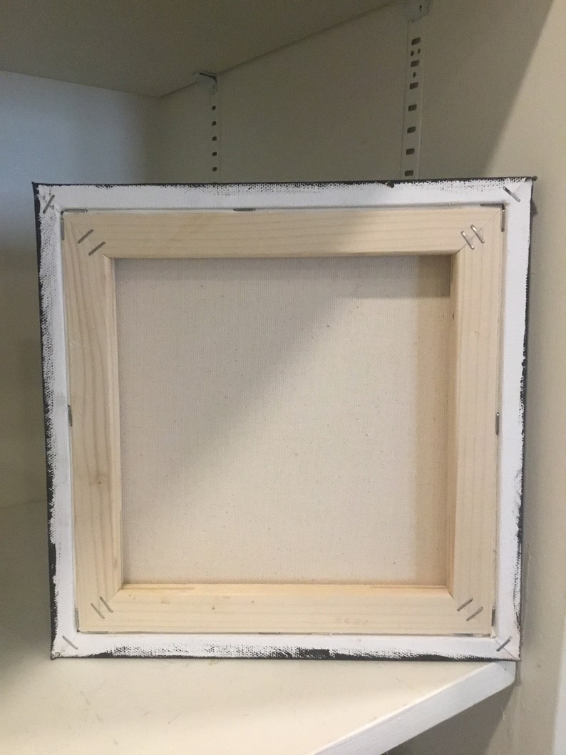 May include: The back of a stretched canvas painting with a wooden frame. The canvas is white and the frame is painted black with white edges.