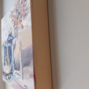 May include: A painting of a still life with two glass bottles and a bouquet of pink flowers. The bottles are clear and have a blue tint. The flowers are arranged in a vase. The painting is on a canvas with a light brown frame.