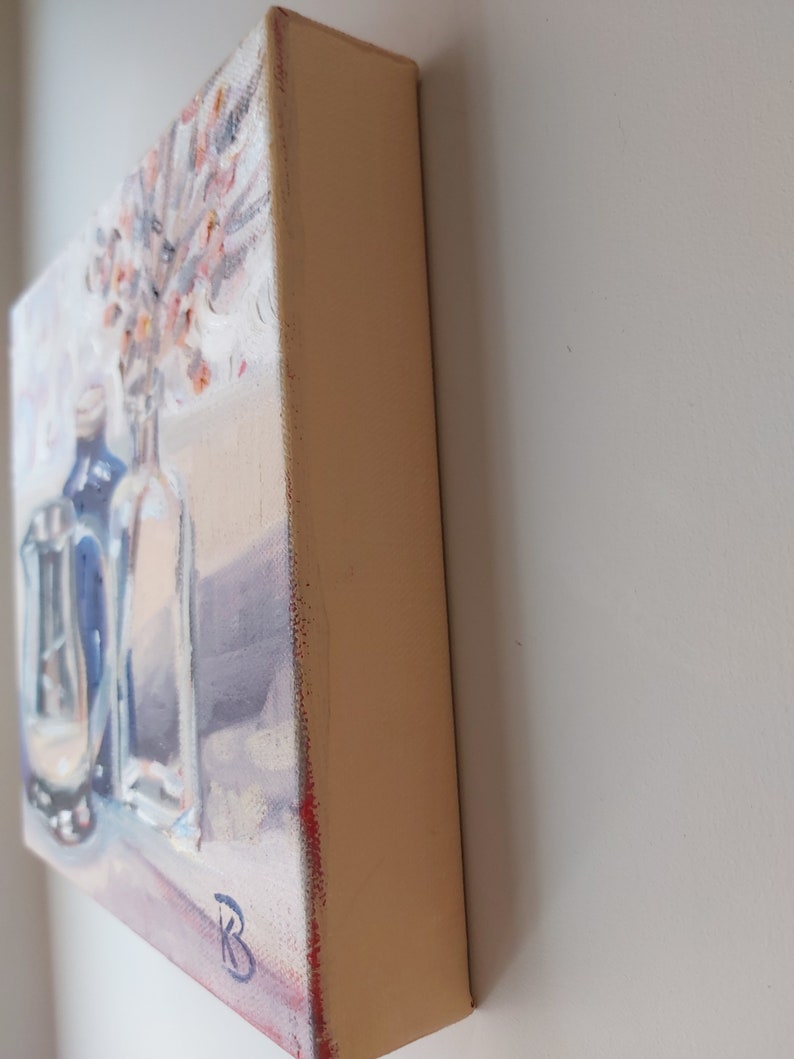 May include: A painting of a still life with two glass bottles and a bouquet of pink flowers. The bottles are clear and have a blue tint. The flowers are arranged in a vase. The painting is on a canvas with a light brown frame.