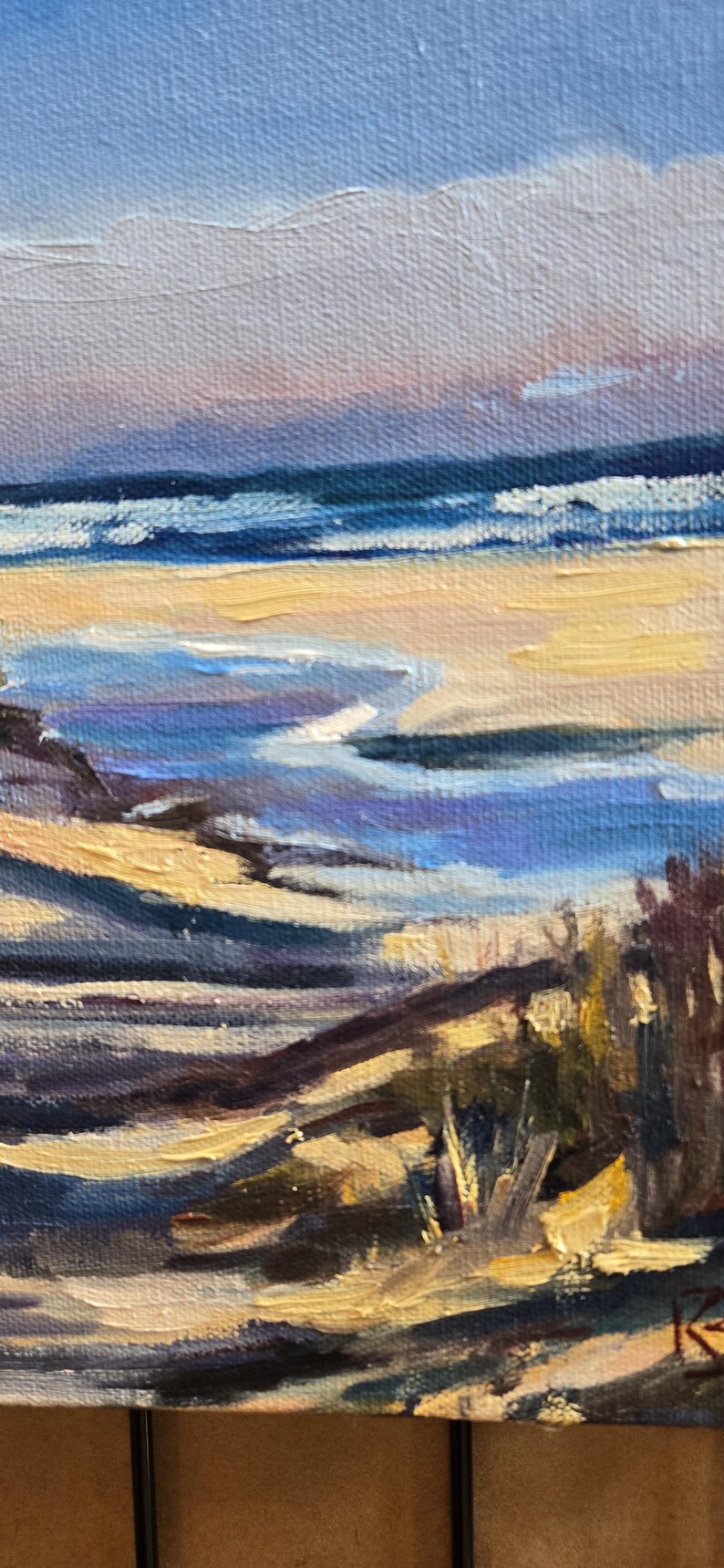 May include: A painting of a beach scene with blue water, yellow sand, and purple and brown hills in the foreground. The sky is a light blue with a pink hue.
