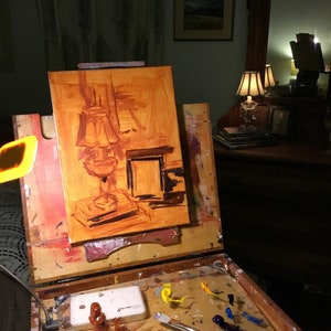 May include: A wooden easel with an oil painting in progress. The painting depicts a still life of a lamp with a brown shade and a book. The easel has a palette with various colors of paint and a paintbrush.