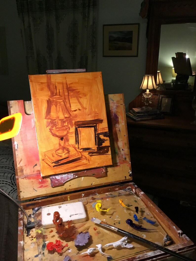 May include: A wooden easel with an oil painting in progress. The painting depicts a still life of a lamp with a brown shade and a book. The easel has a palette with various colors of paint and a paintbrush.