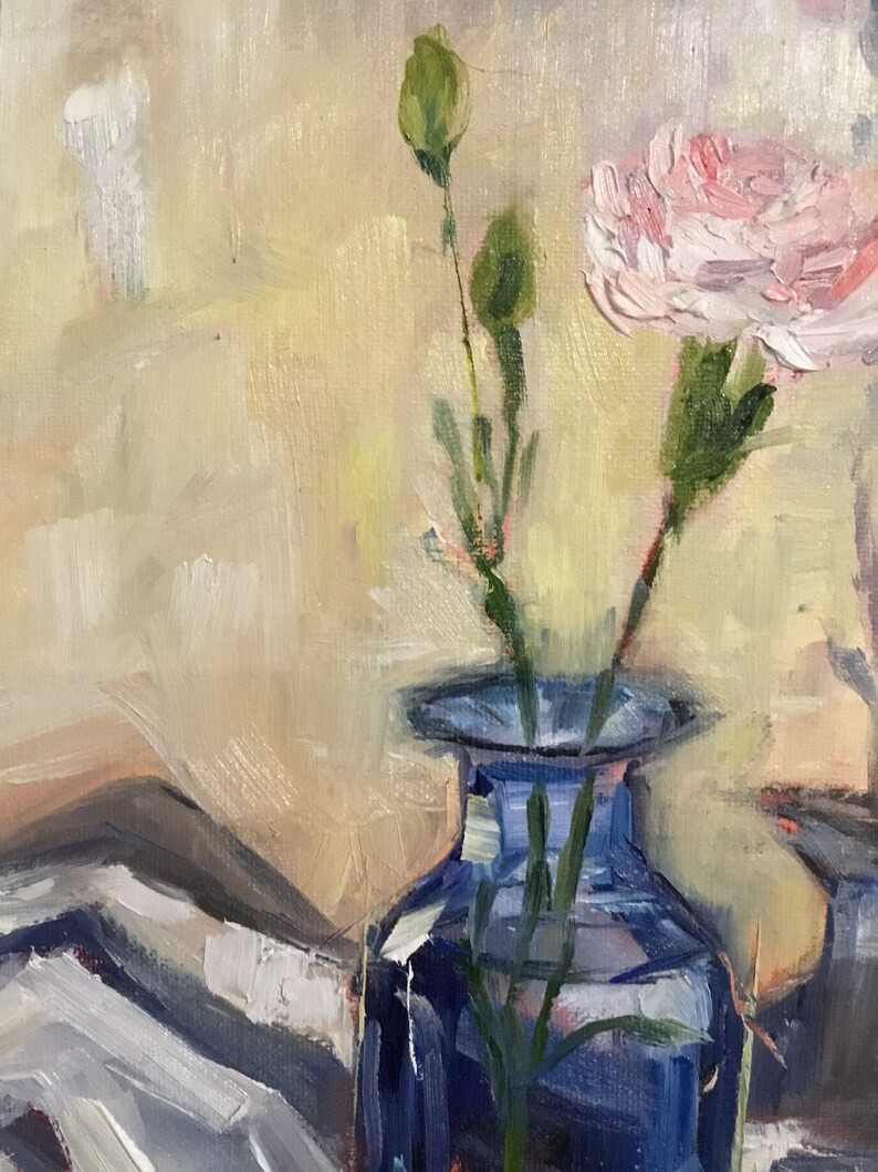 May include: An oil painting of a blue glass vase holding a pink carnation and two green buds. The background is a soft blend of yellow and white, with visible brushstrokes. A white and gray patterned cloth is visible in the lower left corner.