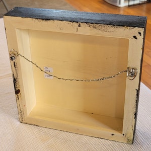 May include: The back of a framed artwork with a black frame and a white mat. A thin wire is attached to the frame with two metal hooks. A handwritten label on the back of the frame reads "48 054 150".