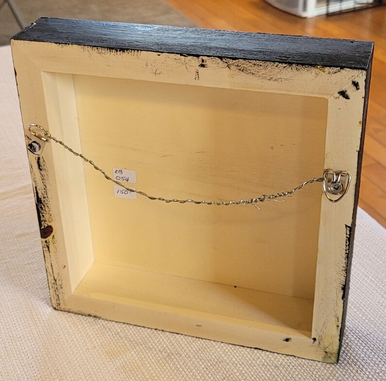 May include: The back of a framed artwork with a black frame and a white mat. A thin wire is attached to the frame with two metal hooks. A handwritten label on the back of the frame reads "48 054 150".