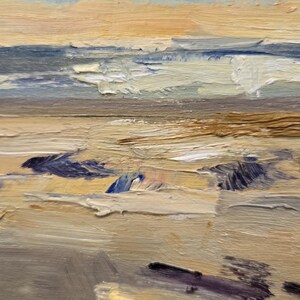 May include: An oil painting on canvas depicting a coastal scene with thick, textured brushstrokes. The composition features a sandy beach in the foreground with dark purple shadows, transitioning to blue ocean waves and a soft, cloudy sky in shades of peach.