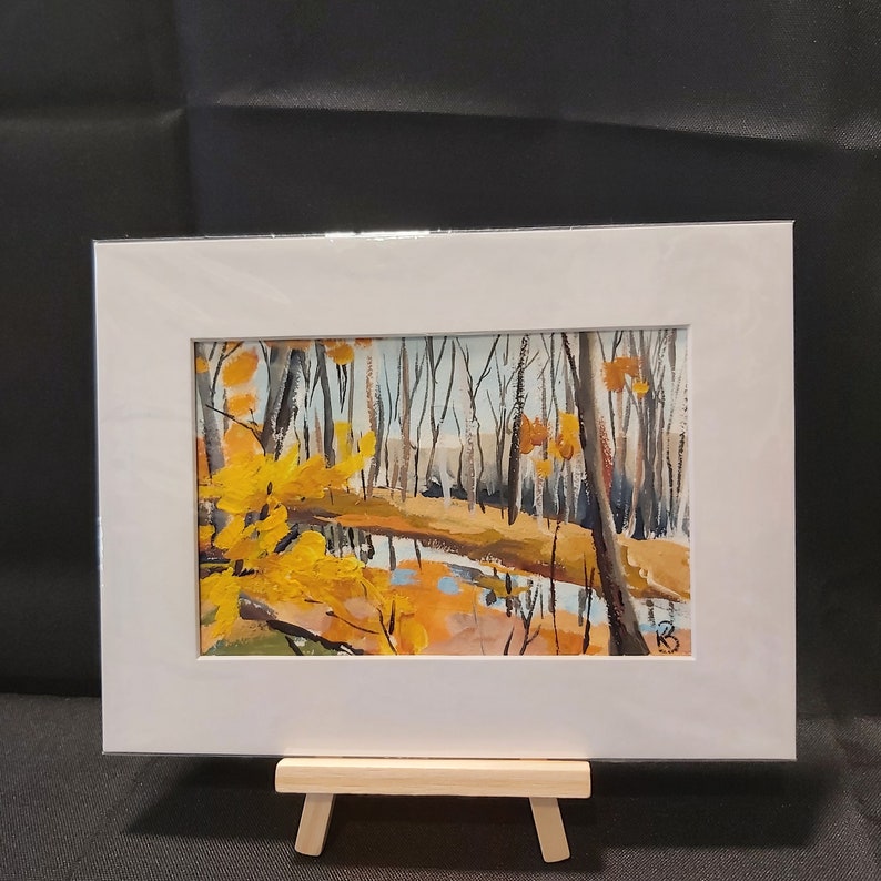 May include: A framed painting of a forest scene with a stream running through it. The trees are bare and the leaves are a vibrant yellow. The painting is set against a white background.