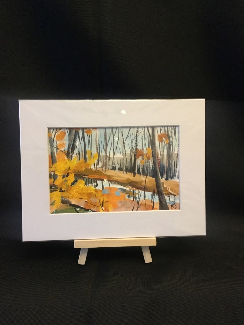 May include: A framed painting of a forest scene with a stream running through it. The trees are mostly bare, but some have yellow leaves. The stream is a light blue color and reflects the sky. The painting is on a small easel.