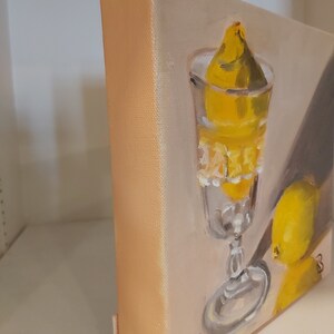 Op de afbeelding: Olieverfschilderij van een stilleven met een glas met een citroen erin en een andere citroen ernaast. Het kunstwerk gebruikt een warm kleurenpalet van geel en beige. Het doek is op een houten frame gemonteerd.