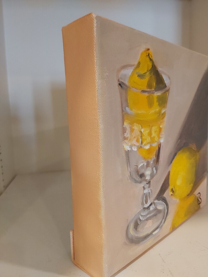 Op de afbeelding: Olieverfschilderij van een stilleven met een glas met een citroen erin en een andere citroen ernaast. Het kunstwerk gebruikt een warm kleurenpalet van geel en beige. Het doek is op een houten frame gemonteerd.
